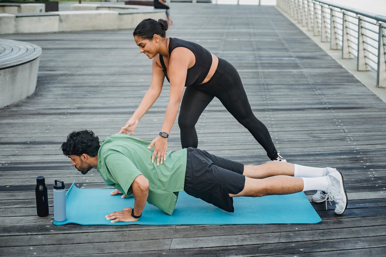 hero-img Man doing push-ups with assistance from a trainer on a boardwalk during summer.