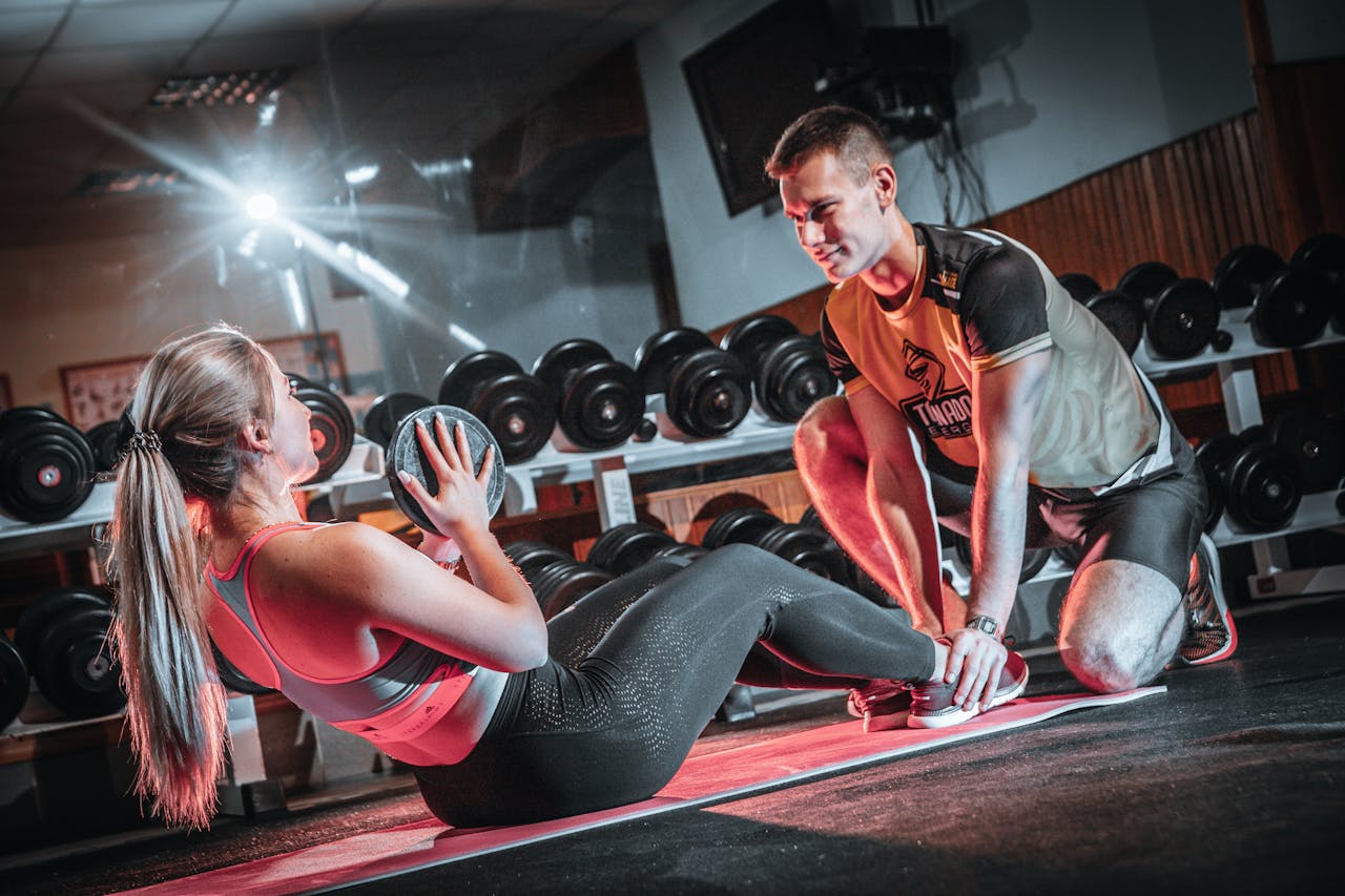 about-01 A personal trainer assists a woman doing sit-ups with a medicine ball in a gym setting.