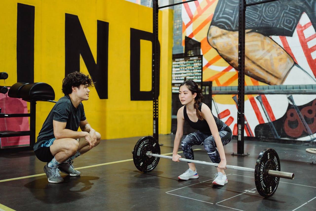services-01 A young woman lifting weights guided by a trainer in a colorful gym setting.